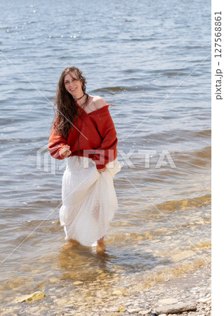 A young woman with long hair and blue eyes, in a white skirt and a terracotta sweater, runs barefoot on the water against the backdrop of the sea and laughs 127568861