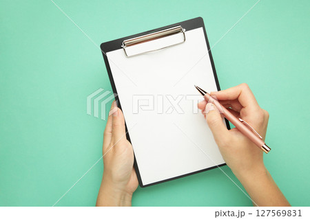 A female hand hold a pen and black clipboard with paper on mint background. A female hand hold a pen and black clipboard with paper on mint background. 127569831
