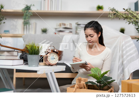 Moving and Organizing. Young woman writing notes while preparing for relocation in a cozy living space. 127572613