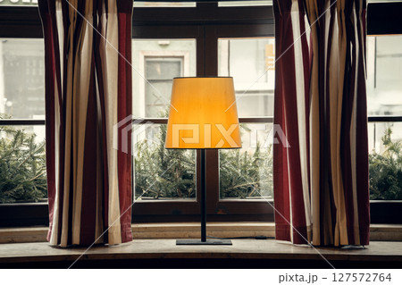 A warmly lit table lamp on a rustic windowsill, surrounded by striped curtains and greenery outside A warmly lit table lamp on a rustic windowsill, surrounded by striped curtains and greenery outside 127572764