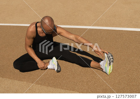 Athlete wearing black sleeveless tank top stretching legs after workout, warming up at sport stadium . Outdoors training. Morning routine. Healthy and sport lifestyle concept. 127573007