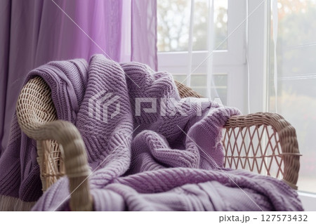 Serene minimalist living room featuring a plush armchair draped with a purple knitted throw blanket, soft natural light and clean-lined furniture in delicate soft tones 127573432