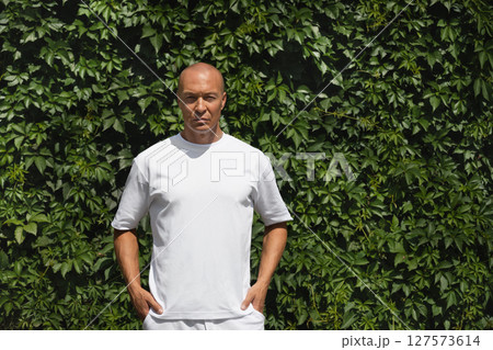 Outdoor portrait of serious caucasian adult man wearing white t-shirt against plant wall of green leaves. Eco concept. Mock up. Sustainable lifestyle. Outdoor portrait of serious caucasian adult man wearing white t-shirt against plant wall of green leaves. Eco concept. Mock up. Sustainable lifestyle. 127573614
