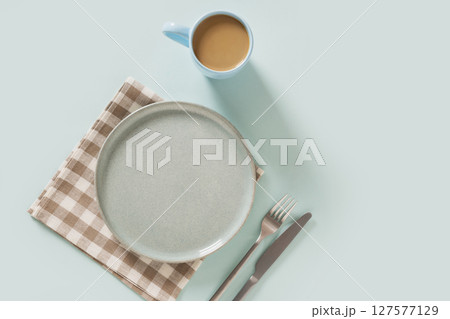 Empty ceramic blue plate, coffee and checkered napkin on plain light blue background. Table setting for breakfast. Mock up. View from above. Copy space Empty ceramic blue plate, coffee and checkered napkin on plain light blue background. Table setting for breakfast. Mock up. View from above. Copy space 127577129