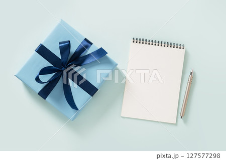 Blue gift box, white blank, spiral notebook and pen on pastel blue background. Invitation. Flat lay. View from above. Copy space. 127577298