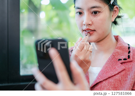 A young woman applies lipstick while using her smartphone, set against a bright, modern background. 127581066
