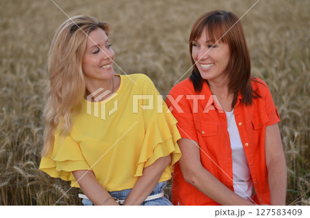 Two women enjoying a joyful moment in a golden wheat field during sunset while wearing bright summer outfits in a rural setting 127583409