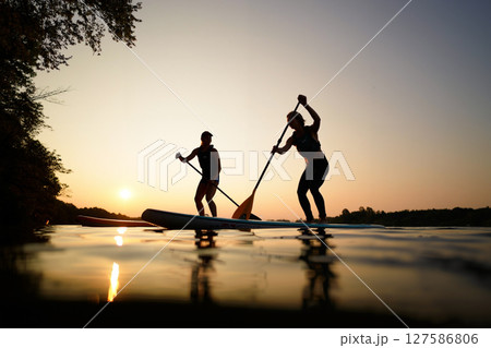 Paddleboarders Gliding Across Calm Water at Sunset With Orange and Purple Skies 127586806