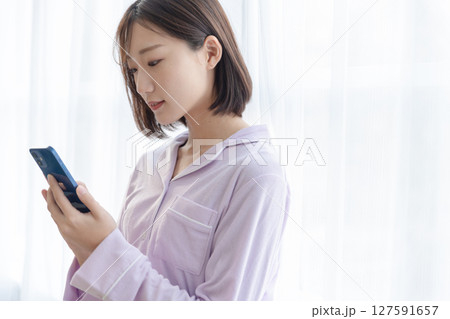 窓辺でパジャマを着てスマホを操作している若い女性 127591657