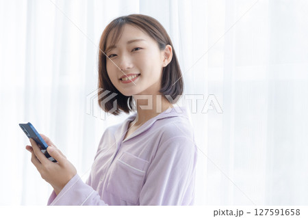 窓辺でパジャマを着てスマホを操作している若い女性 127591658