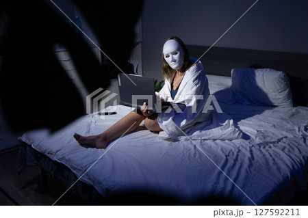 User Wearing a Mask Sits on Bed With Laptop in Dark Room Highlighting Digital Privacy Concerns User Wearing a Mask Sits on Bed With Laptop in Dark Room Highlighting Digital Privacy Concerns 127592211