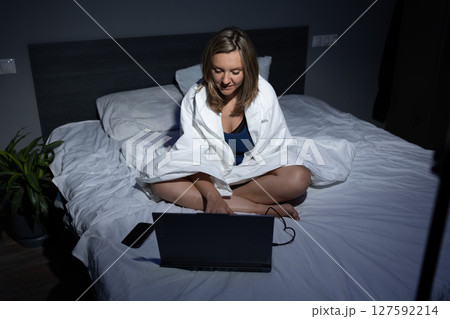 Woman Working on Laptop in Bed While Wrapped in a Blanket in a Cozy Bedroom at Night 127592214