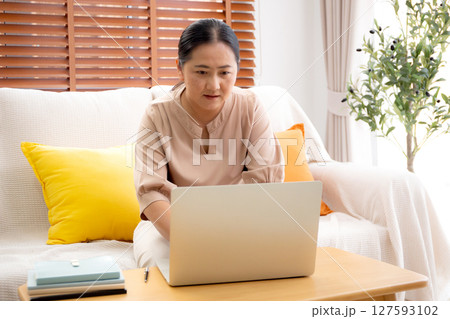 Asian woman working or studying online using a laptop sitting on sofa in living room at home. Asian woman working or studying online using a laptop sitting on sofa in living room at home. 127593102