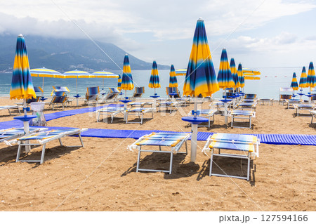 Sunny beach with colorful umbrellas and lounge chairs by the Adriatic Sea. Summer relaxation, coastal tourism and Mediterranean leisure in Montenegro. 127594166