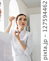 Confident female cosmetologist holding facial serum with dropper in front of mirror, wearing white coat, promoting skincare product for healthy radiant skin, anti-aging routine in beauty clinic 127594462