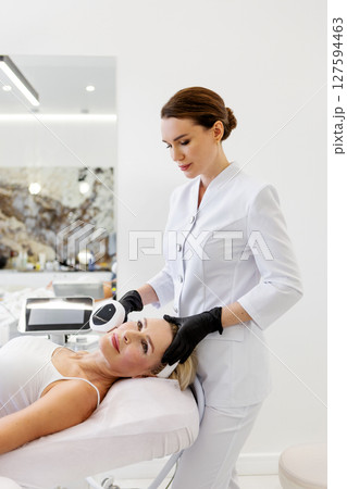 Mature woman receiving anti-aging skin treatment lying in cosmetic clinic. Cosmetologist performing photorejuvenation face procedure using professional aesthetic laser machine, skincare and wellness 127594463