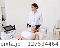 Professional female cosmetologist performing facial skin treatment using modern equipment in luxury beauty clinic, mature woman receiving rejuvenation therapy lying on chair in clean white interior 127594464