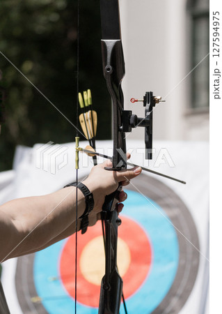 Archer Aiming Recurve Bow at Target Outdoors: Focused Archery Practice with Arrows, Colorful Target, Green Foliage, Black Equipment Archer Aiming Recurve Bow at Target Outdoors: Focused Archery Practice with Arrows, Colorful Target, Green Foliage, Black Equipment 127594975