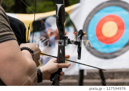 Archer Aiming Compound Bow at Target: Outdoor Archery Sport with Blue Target and Focused Male Participant, Black Equipment and Natural Light 127594976