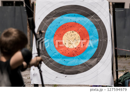 Archery Practice at Outdoor Range: Boy Shooting Arrows at Bullseye Target with Scattered Impacts, Wooden Stand, Blue and Brown Colors Archery Practice at Outdoor Range: Boy Shooting Arrows at Bullseye Target with Scattered Impacts, Wooden Stand, Blue and Brown Colors 127594979