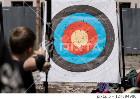 Boy Archery Practice Outdoors: Target Board, Arrows, Backyard, Summer Sports Activity, Blue, Red, Black, Training, Skill Development, Competition. 127594980