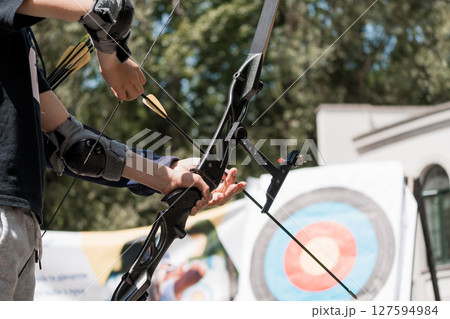 Archery Practice in Sunlight: Close-Up of Archer Drawing Bow with Arrows Aimed at Target Outdoors, Blue Shirt, Focused Action Archery Practice in Sunlight: Close-Up of Archer Drawing Bow with Arrows Aimed at Target Outdoors, Blue Shirt, Focused Action 127594984