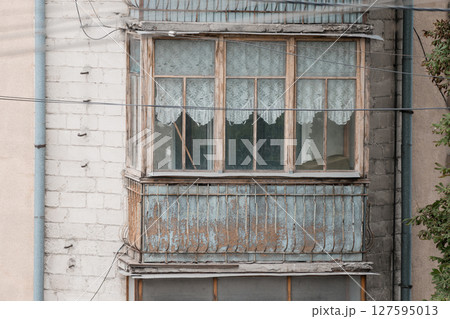 Dilapidated Balcony Window in Eastern Europe: Aged Wood, Lace Curtains, Concrete Building Facade, Rusty Metal Railing, Vintage Aesthetic. 127595013