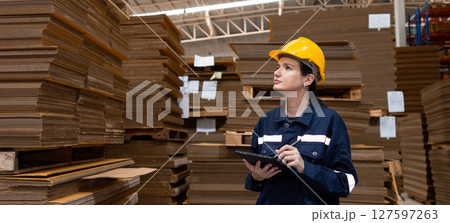 woman paper warehouse worker using tablet checking inspecting quality cardboard carton in storage. Large industry in paper product line manufacturing. Employee level wear uniform and helmet yellow. 127597263