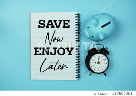 Save Now Enjoy Later text on notebook paper and piggy bank and alarm clock on blue background Save Now Enjoy Later text on notebook paper and piggy bank and alarm clock on blue background 127605361