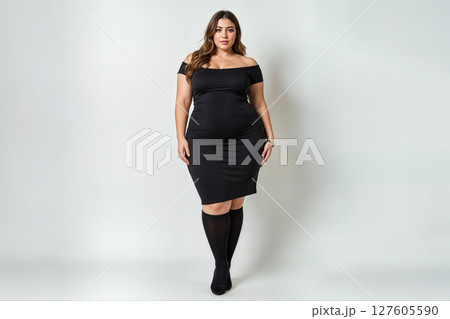 Confident plus-size woman in a black off-the-shoulder dress and knee-high socks. 127605590