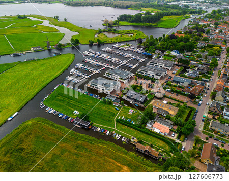 A stunning aerial view of a marina surrounded by green fields, waterways, boats, and a nearby residential area, with a windmill and open landscape in the distance. 127606773