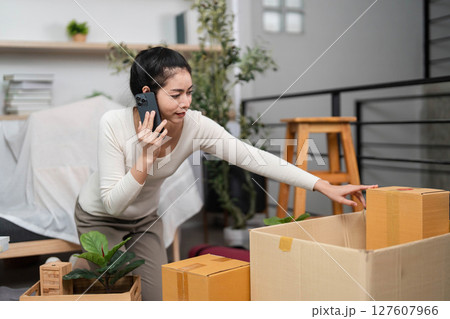 Moving and Communication. Young woman talking on phone while organizing boxes for relocation. Moving and Communication. Young woman talking on phone while organizing boxes for relocation. 127607966