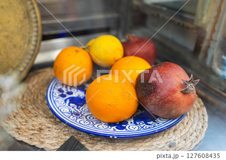 Citrus and pomegranate fruits rest on a traditional blue patterned plate. Mediterranean freshness, market display, and rustic natural nutrition. Citrus and pomegranate fruits rest on a traditional blue patterned plate. Mediterranean freshness, market display, and rustic natural nutrition. 127608435