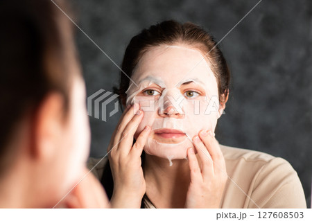 Late night skincare routine. Woman applying moisturizing face mask. Reflection in bathroom mirror. Dermatology concept. 127608503