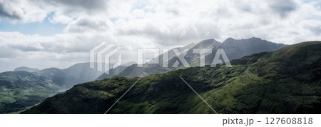 A scenic view showing the rolling green hillsides and imposing mountains of the Gap of Dunloe under a sky filled with dramatic clouds. Rain shafts are visible in the distance. 127608818