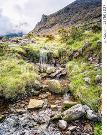 A small waterfall cascades over rocks and vegetation in a picturesque landscape. This is along the popular hiking trail of Carrauntoohill in County Kerry, Ireland, with a mountain in the background. 127608837