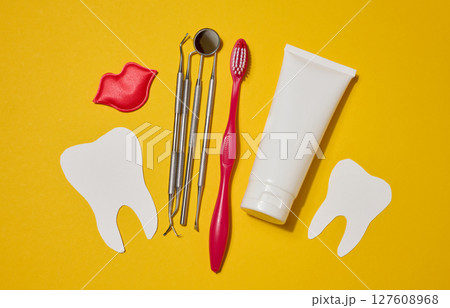Red toothbrush, white tube of toothpaste, red decorative lips, dental instruments, dental floss in a case and white paper teeth cut out on a yellow background. 127608968