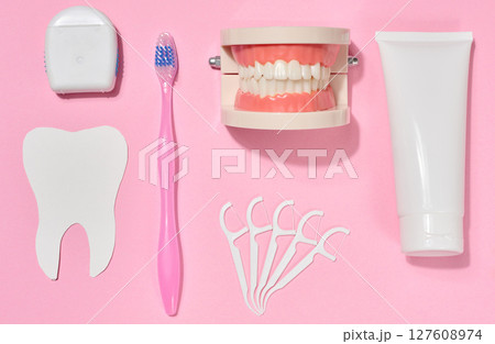 White tube of toothpaste, pink toothbrush, dental floss, plastic floss holders, denture model on pink background. 127608974
