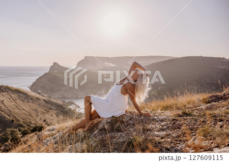 A woman is sitting on a hillside overlooking the ocean. She is wearing a white dress and has blonde hair. The scene is serene and peaceful, with the ocean in the background. 127609115