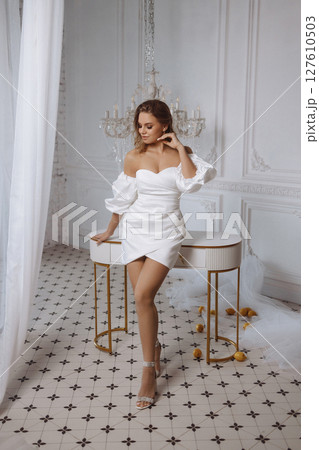 Young woman in white dress posing near table in classic white room with tiled floor and lemons 127610503