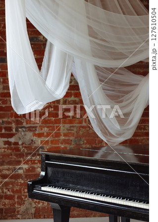 Old black grand piano in loft room with red brick wall and white fabric drapes 127610504