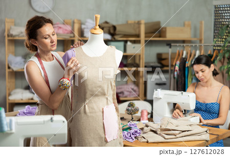 Female tailor sews new dress on mannequin in a sewing studio. Other woman tailor is sewing on sewing machine 127612028
