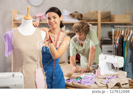 Young female dressmaker working with mannequin while assistant drawing fashion sketch Young female dressmaker working with mannequin while assistant drawing fashion sketch 127612086