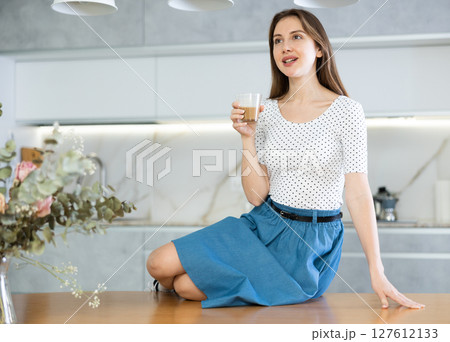 Portrait of cheerful contented woman with cup of cocoa in her hands in modern kitchen Portrait of cheerful contented woman with cup of cocoa in her hands in modern kitchen 127612133