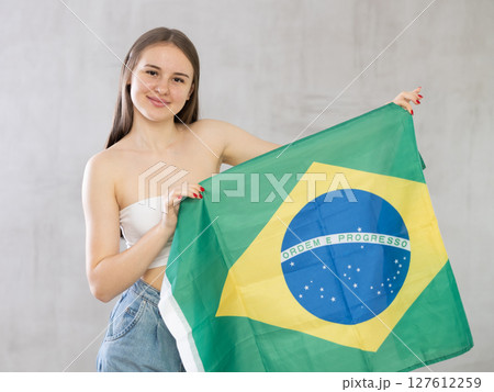 Reliant positive young woman holds big national flag of Brazil with confident smile Reliant positive young woman holds big national flag of Brazil with confident smile 127612259