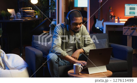 Focused black guy teleworking on laptop in a living room feels happy with task completion, achieving success. Remote worker balancing comfort and work efficiency in a flexible workspace. Camera B. 127613499