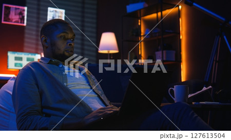 Skilled african american man multitasking on laptop in cool living room, connected to the internet while managing freelance work and remote responsibilities. Browsing web on bean bag. Camera A. Skilled african american man multitasking on laptop in cool living room, connected to the internet while managing freelance work and remote responsibilities. Browsing web on bean bag. Camera A. 127613504