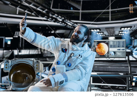 Astronaut wearing spacesuit inside futuristic spacecraft doing selfie with smartphone. Smiling spaceman wearing equipment doing photos with mobile phone during space travel operations Astronaut wearing spacesuit inside futuristic spacecraft doing selfie with smartphone. Smiling spaceman wearing equipment doing photos with mobile phone during space travel operations 127613506