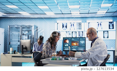 Old specialist meeting with a woman patient to discuss results in a cabinet, using advanced diagnostic equipment to interpret x ray scans and results, professional healthcare approach. Camera A. 127613534