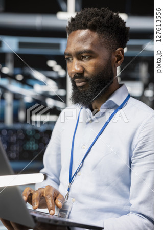 Computer scientist in server room uses laptop to implement and manage data backup solutions. African american man in data center identifying and resolving technical errors, ensuring efficient recovery 127613556